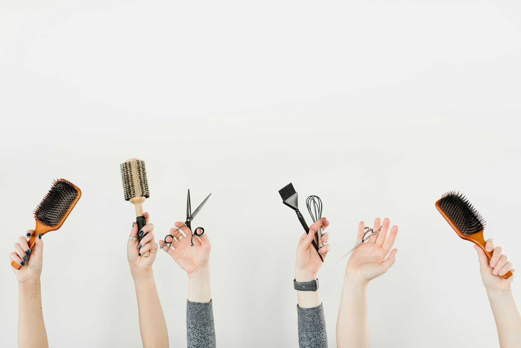 Raised hands holding hairbrushes, scissors and a tint brush against a white wall