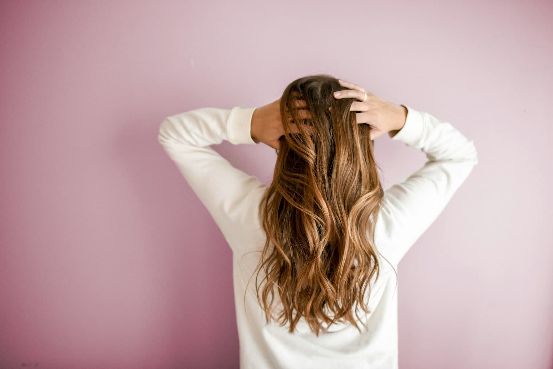 Woman seen from behind with long, wavy brown hair against a pink wall